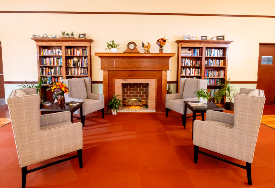 A cozy living room area with four beige armchairs arranged around two small dark wooden tables. The room features a brick fireplace with a wooden mantel decorated with a clock, a rooster figurine, and a vase of flowers. On either side of the fireplace are tall wooden bookshelves filled with books and decorative items. The floor is covered with a red carpet, and there are several potted plants placed around the room.