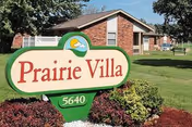 Outdoor view of Prairie Villa facility sign with the address number 5640, set in a landscaped area with bushes and grass, and a brick building in the background under a clear sky.