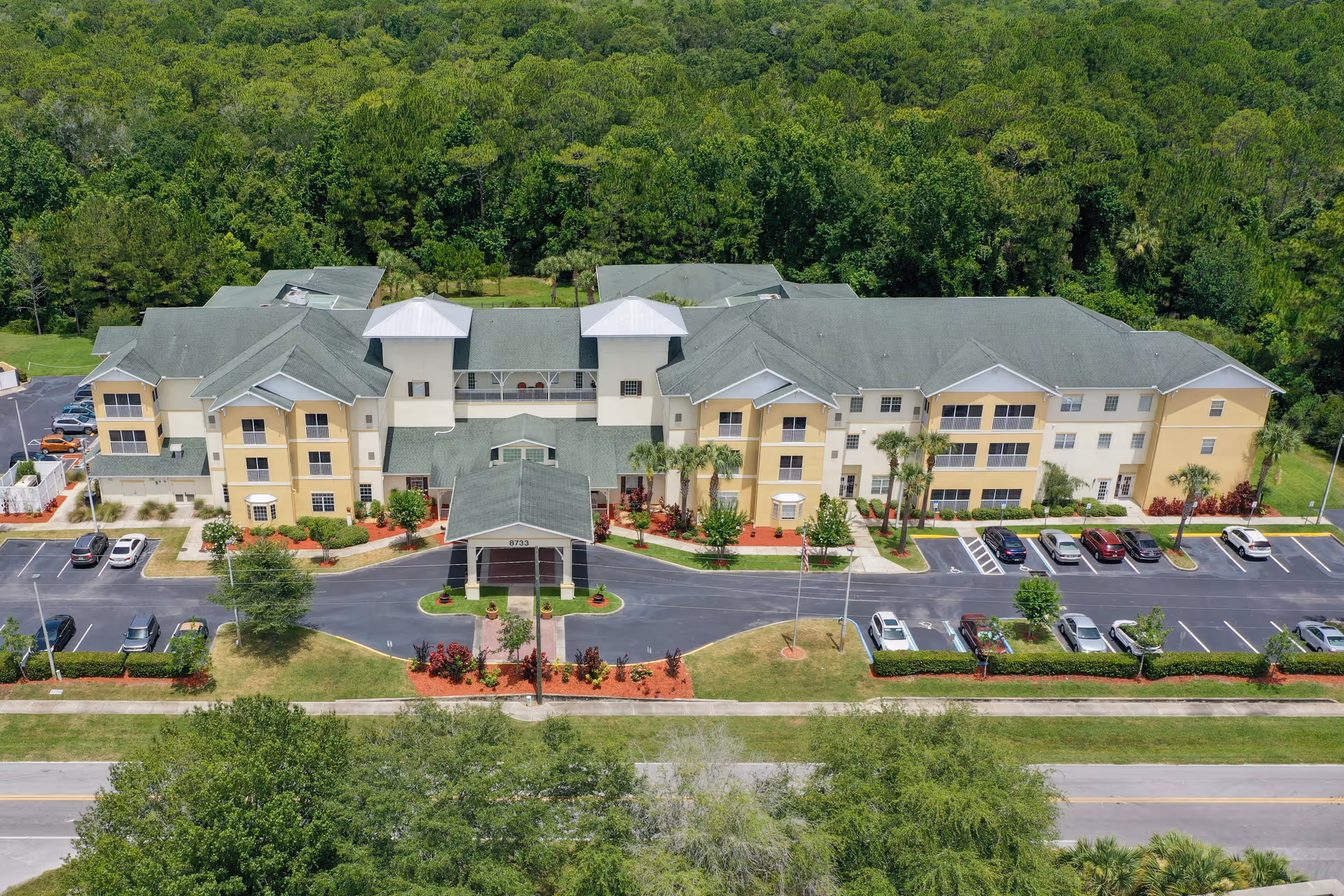 Aerial view of a large, three-story senior living facility named Sunflower Springs surrounded by greenery. The building has a green roof and beige exterior walls with multiple windows and a covered entrance. There are several cars parked in the parking lot in front of the building, and the facility is bordered by trees and landscaped areas.