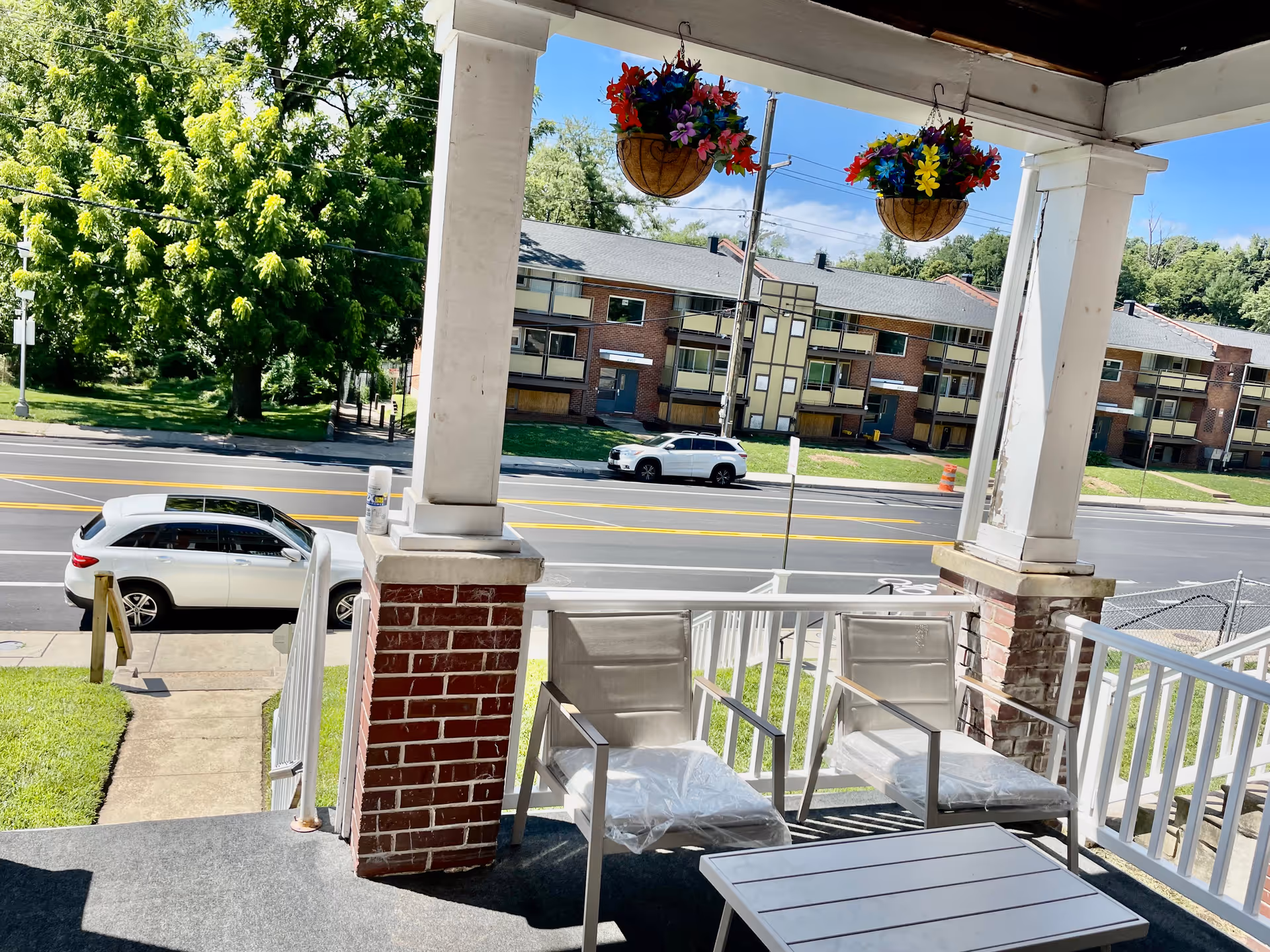 View from a covered porch with two chairs and a small table. Two hanging flower baskets with colorful flowers are suspended from the porch ceiling. Across the street, there are apartment buildings and parked cars. Trees and a clear blue sky are visible in the background.
