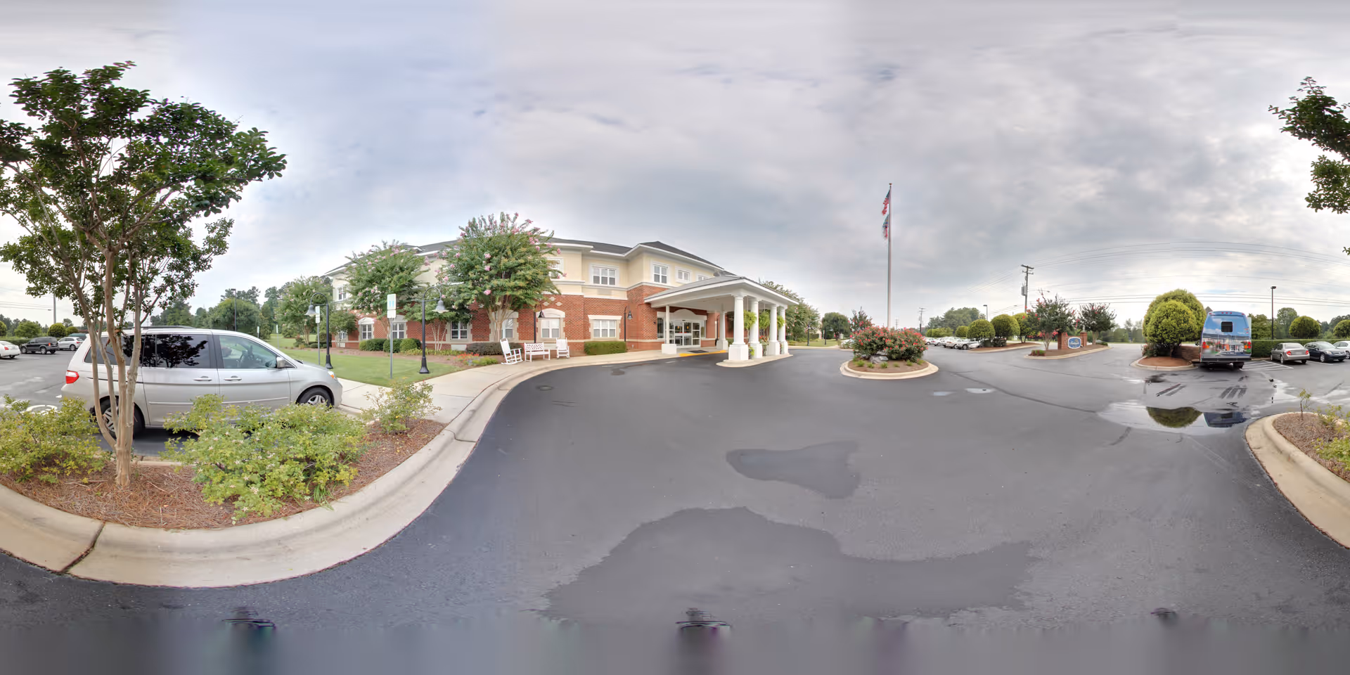 Wide exterior view of Morningside of Gastonia senior living facility showing the front entrance with a covered drop-off area supported by white columns, surrounded by landscaped greenery and trees. Several cars are parked in the parking lot under a cloudy sky.