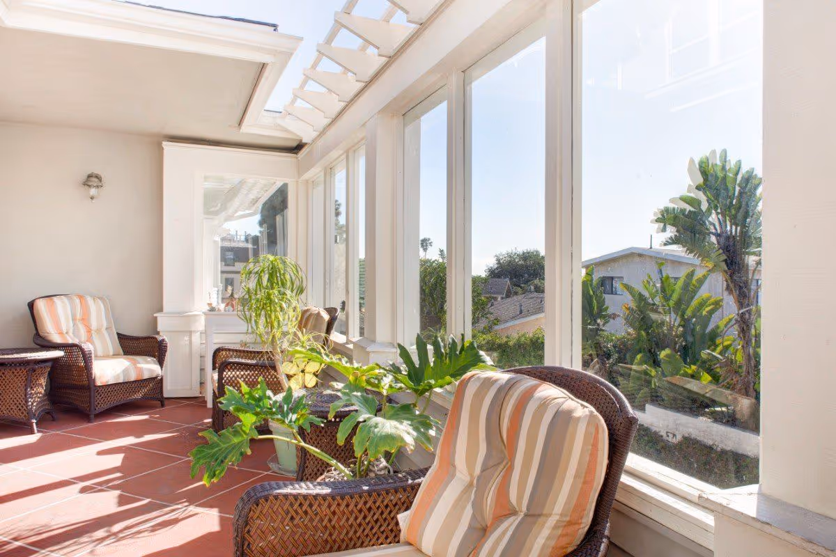 Sunlit enclosed sunroom with wicker chairs, striped cushions, potted plants, and large windows overlooking neighboring houses.