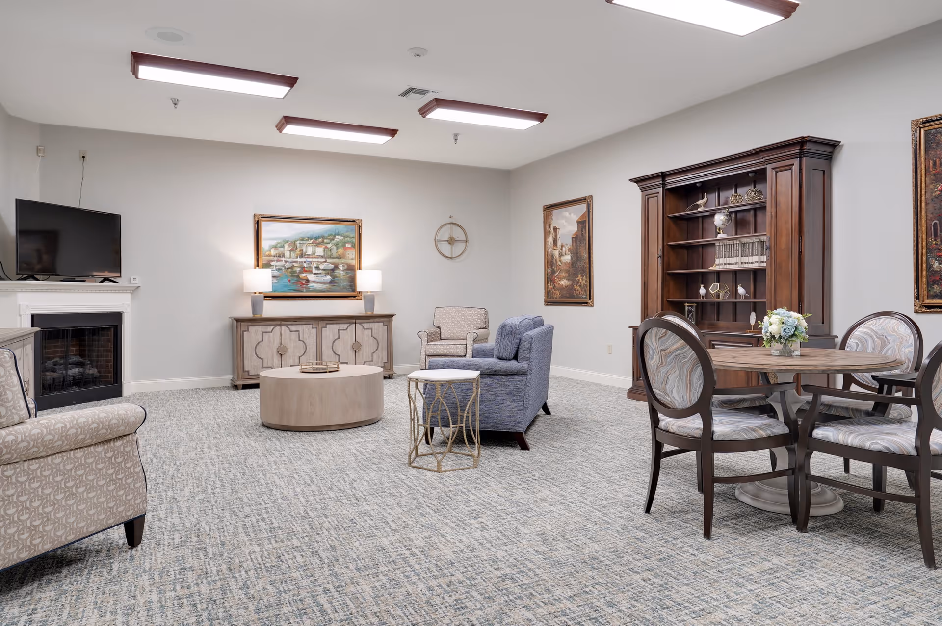 A spacious living room area with patterned carpet, a round wooden coffee table, a blue upholstered sofa, an armchair, and a side table. There is a wooden cabinet with shelves filled with decorative items and books, a round dining table with four chairs, a TV mounted above a white fireplace, and framed paintings on the walls. The room is well-lit with ceiling lights.