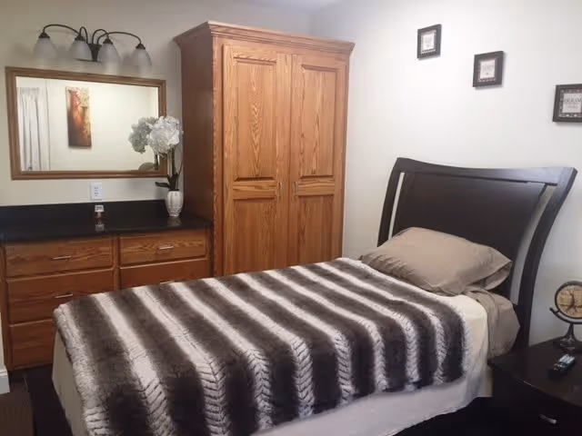 A bedroom with a single bed covered by a striped brown and white blanket. The bed has a brown headboard and beige pillows. Next to the bed is a dark wooden nightstand with a small clock and a remote control on it. Across from the bed is a wooden dresser with a large mirror above it, a vase with white flowers, and a soap dispenser. There is also a wooden wardrobe next to the dresser. The walls are white with three small framed pictures hanging above the bed.
