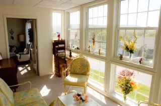 A bright sunroom with large windows overlooking a residential neighborhood. The room contains a floral-patterned chair, a small table with a flower arrangement, a wooden desk with a chair, and various decorative items on the windowsills. Adjacent to the sunroom is a living area with additional seating visible through an open doorway.