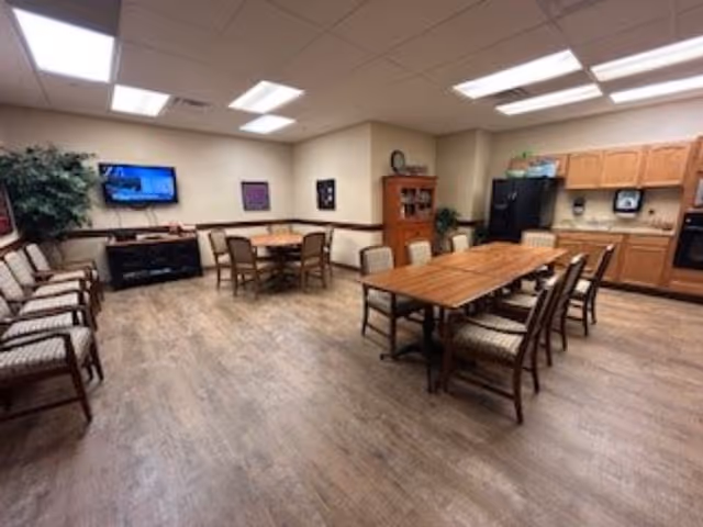 A spacious common room with wooden flooring featuring two wooden tables surrounded by chairs. One table is rectangular with eight chairs, and the other is round with four chairs. The room has a wall-mounted TV, a wooden cabinet, kitchen cabinets with a black refrigerator, and a paper towel dispenser. There are also several chairs lined up against the wall and a potted plant in the corner.