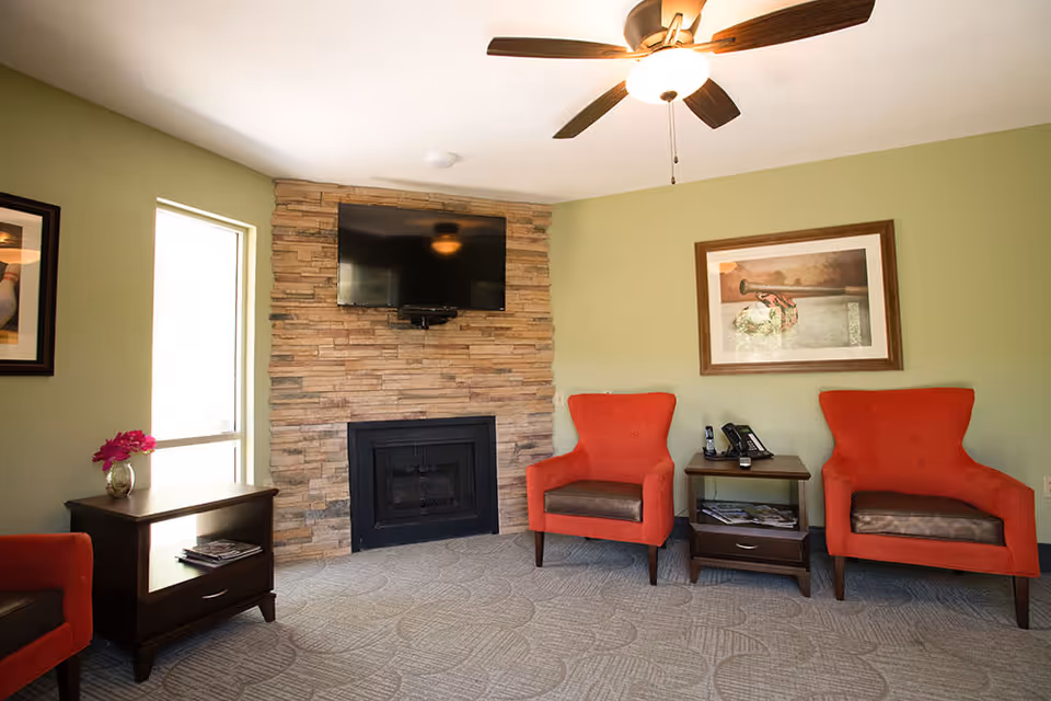 A cozy living room area with two orange armchairs, a small wooden side table with a phone and magazines, a stone fireplace with a mounted flat-screen TV above it, a ceiling fan with lights, and a window letting in natural light.