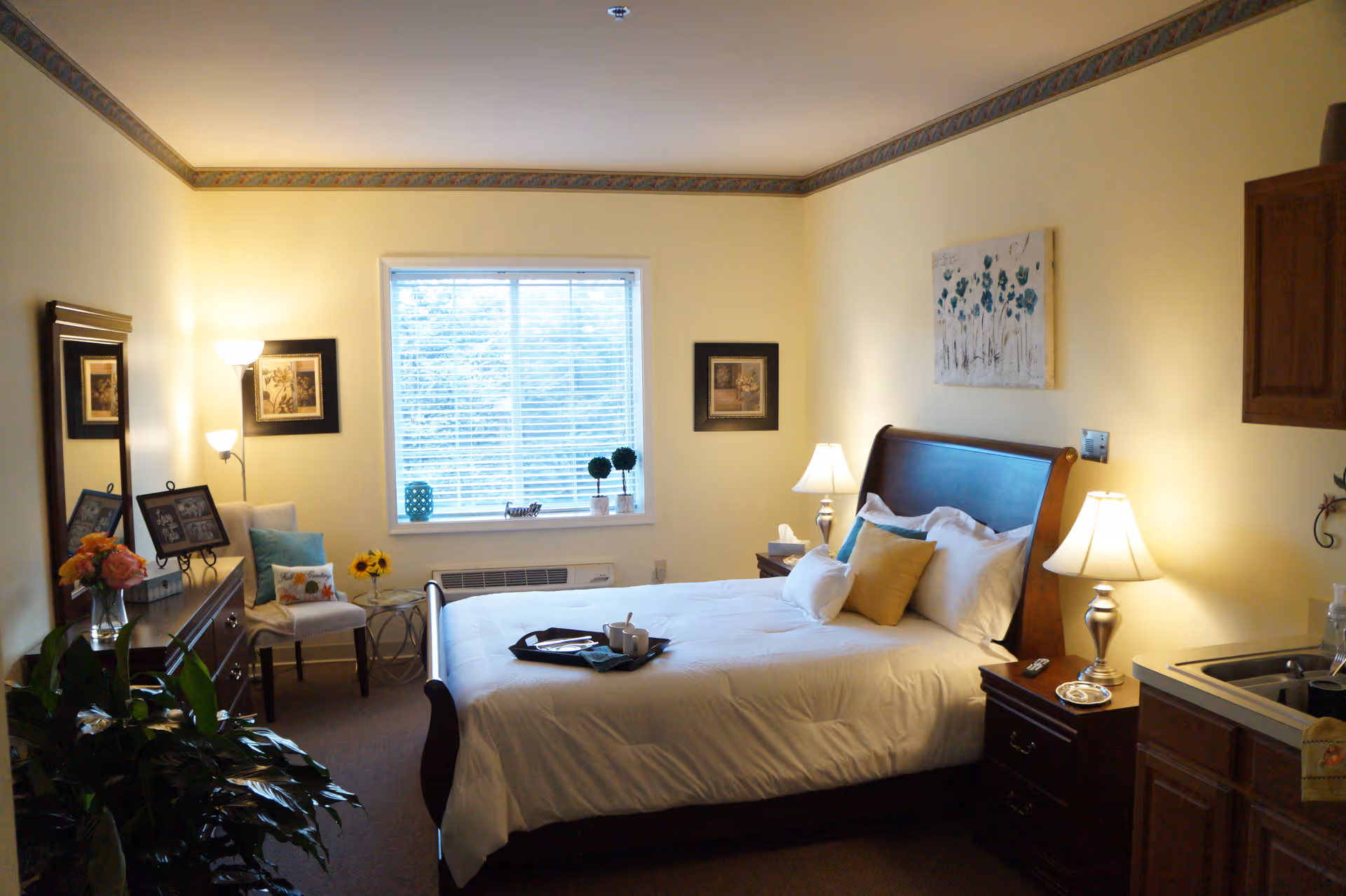 A cozy senior living bedroom with a large bed made with white linens and multiple pillows. The room features a wooden dresser with a mirror, a chair with cushions, two bedside tables each with a lamp, and a window with blinds letting in natural light. There is a small kitchenette area with a sink and cabinets on the right side. The walls are painted light yellow with decorative border trim near the ceiling, and the room is decorated with framed pictures and plants.