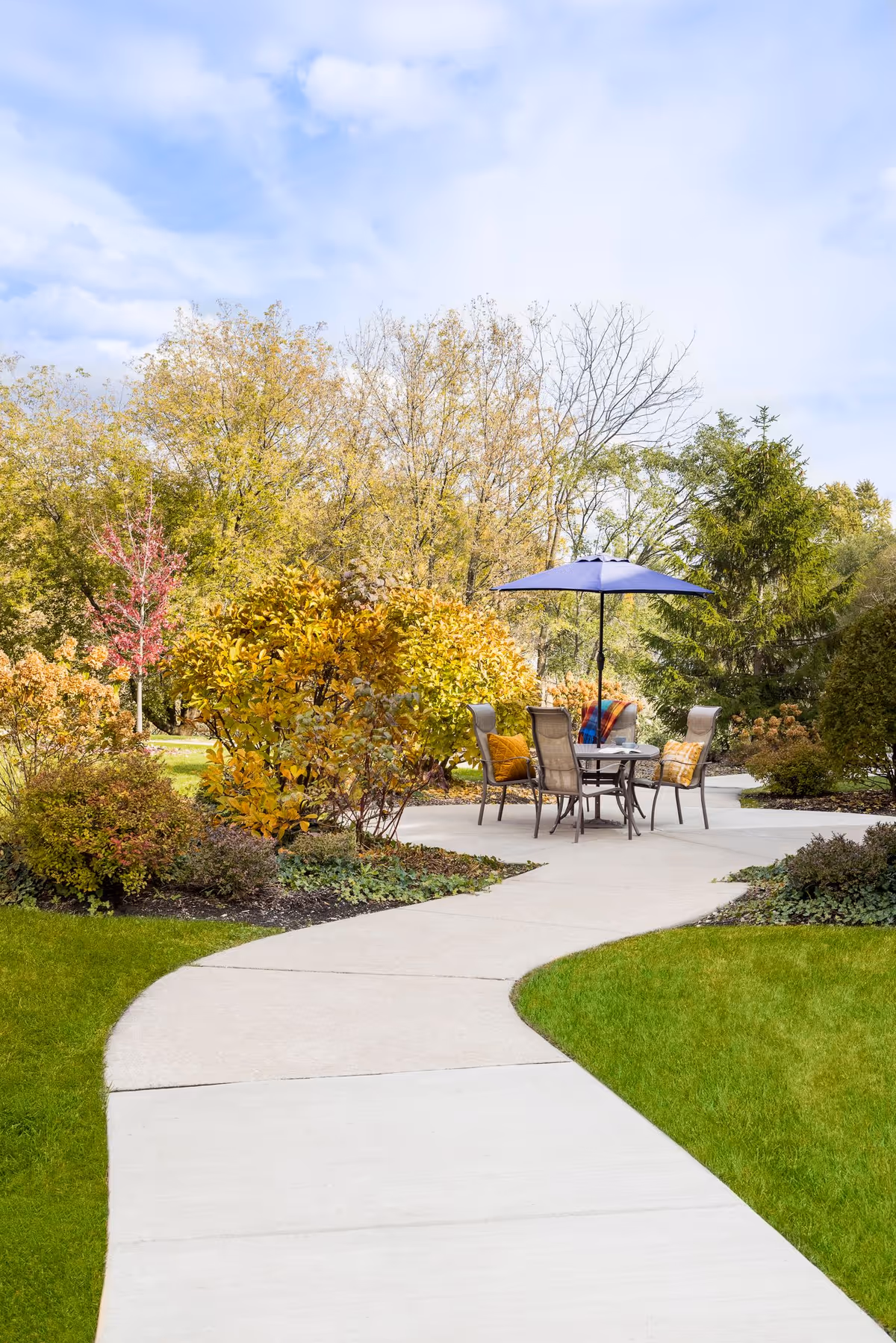 A curved concrete pathway leads to an outdoor seating area with a table, four chairs, and a blue umbrella. The area is surrounded by green grass, bushes, and trees with autumn foliage under a partly cloudy sky.