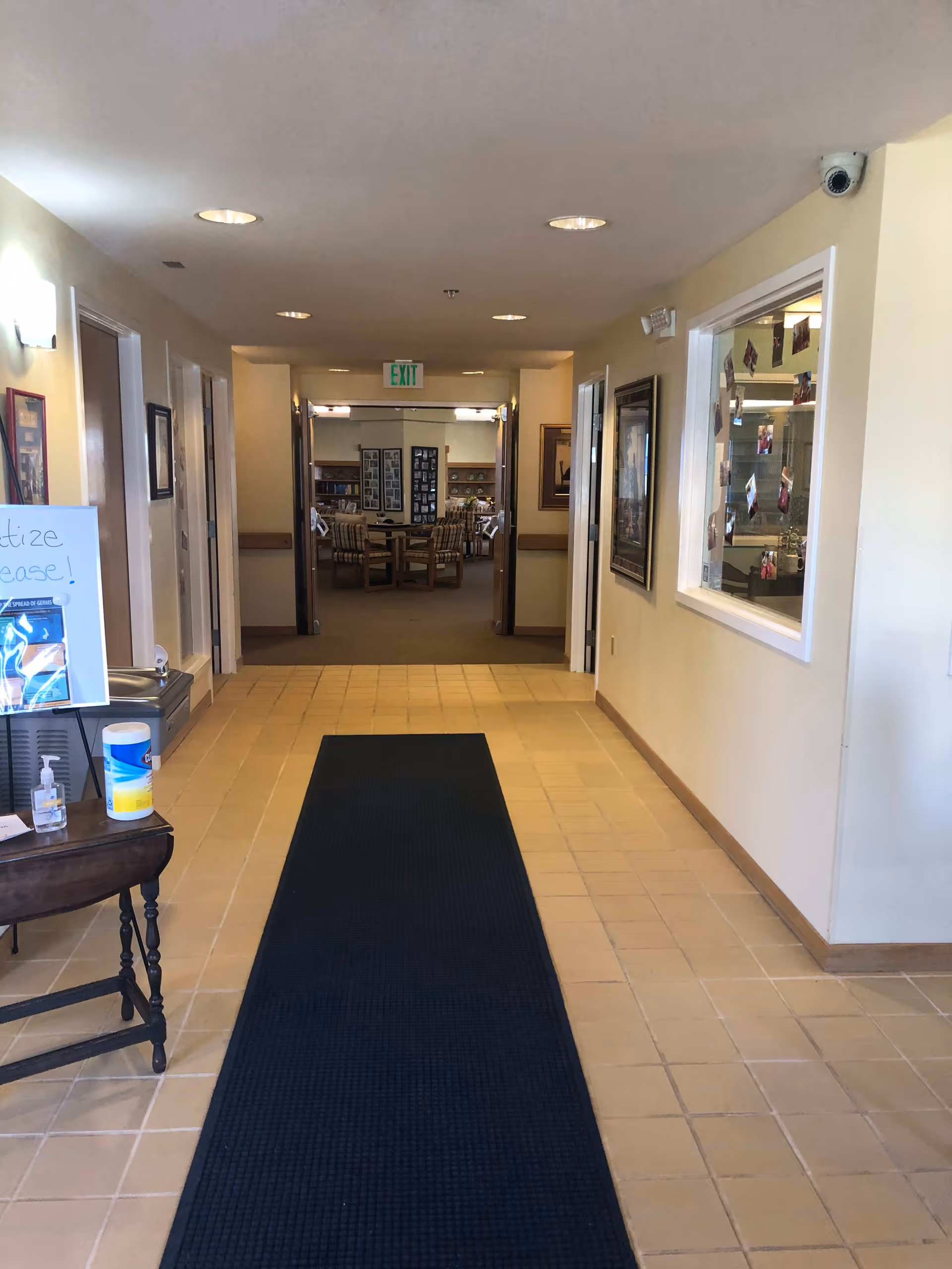 A hallway in a senior living facility with beige tiled floors and cream-colored walls. A dark blue runner rug extends down the center of the hallway. On the left side, there is a small table with hand sanitizer, disinfecting wipes, and a sign encouraging sanitization. The hallway leads to a room with several chairs and tables, visible through double doors. There is an exit sign above the doors and framed pictures on the walls.