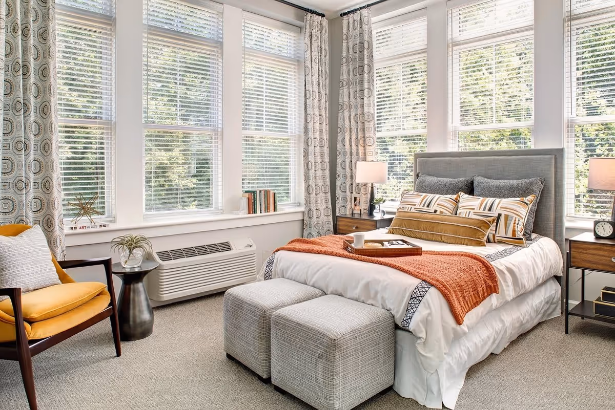 A bright and cozy bedroom with large windows covered by white blinds and patterned curtains. The room features a neatly made bed with multiple pillows and an orange throw blanket, two small ottomans at the foot of the bed, a yellow armchair with a white pillow, a small round side table with a plant, and two wooden nightstands each with a lamp and decorative items.