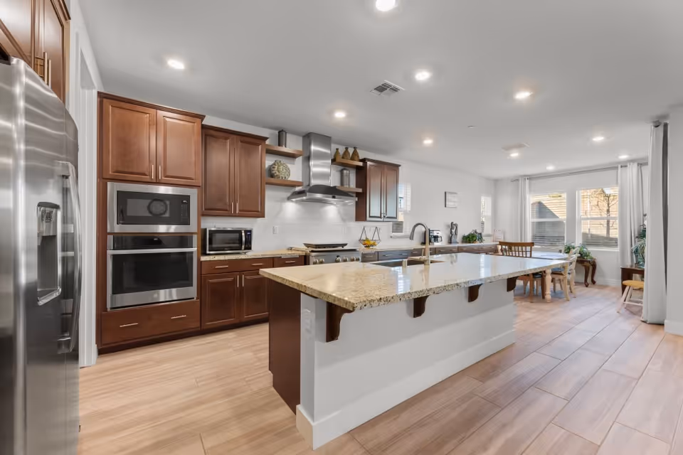 A modern kitchen with wooden cabinets, stainless steel appliances including a refrigerator, microwave, and oven. There is a large granite island countertop with a built-in sink and faucet. The kitchen opens into a dining area with a wooden dining table and chairs near large windows with white curtains, allowing natural light to fill the space. The floor is covered with light wood-style tiles and the ceiling has recessed lighting.