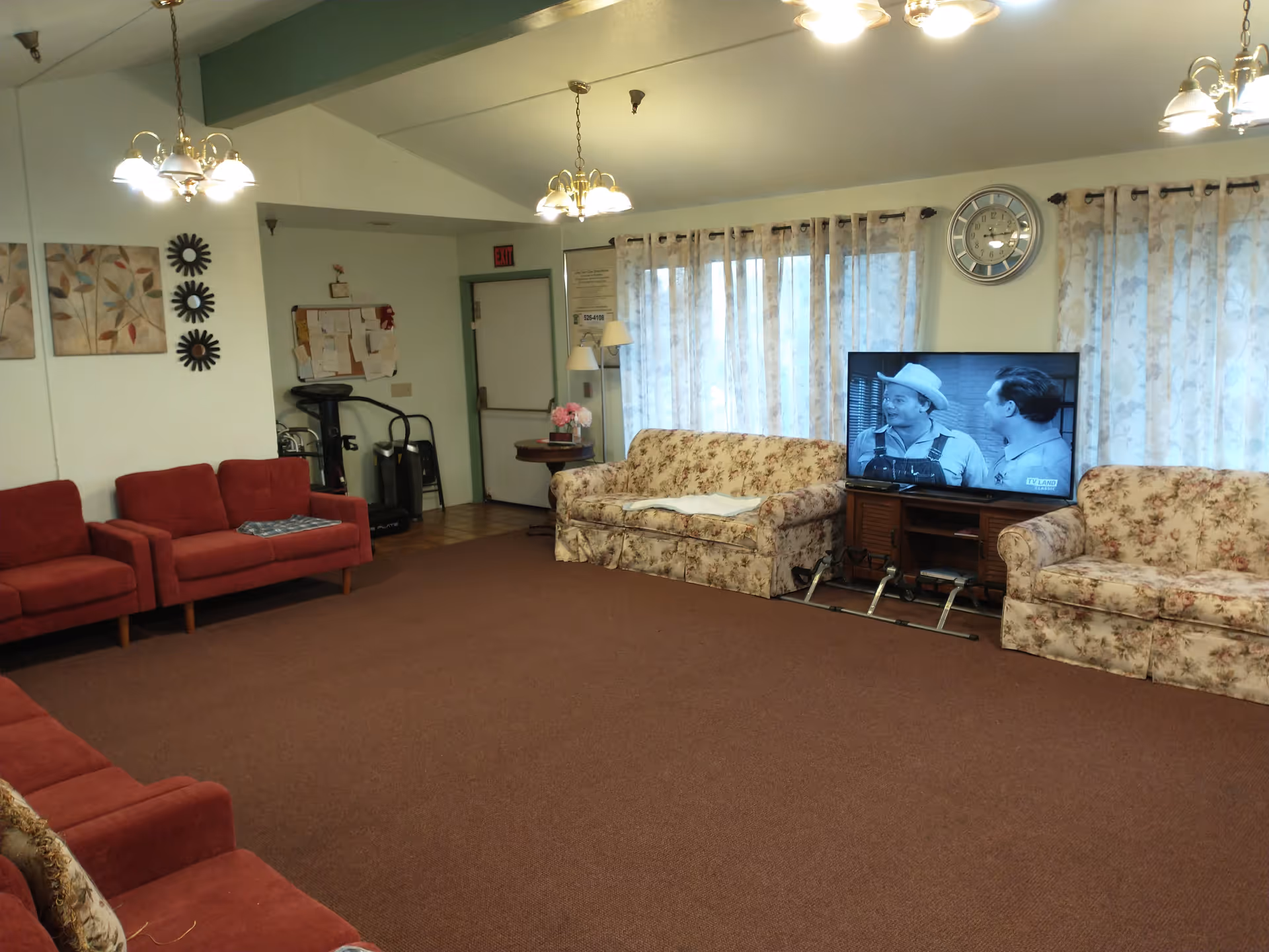 Carpeted common room with red and floral sofas arranged around a TV playing a black-and-white show and wall clock.