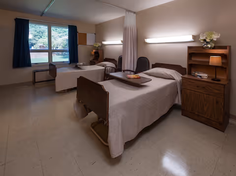A room with two hospital-style beds separated by a privacy curtain. Each bed has a wooden headboard and a bedside table with a lamp and flowers. There is a window with dark curtains letting in natural light, and the floor is tiled.