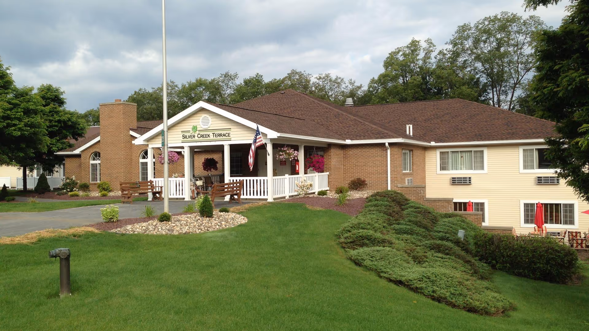 Exterior view of Silver Creek Terrace, a single-story building with a brown roof and brick and beige siding. The entrance features a covered porch with white railings, benches, hanging flower baskets, and an American flag. The surrounding area includes a well-maintained green lawn, shrubs, and trees under a cloudy sky.