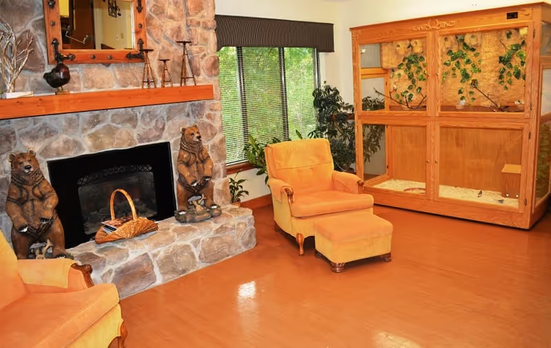 Cozy sitting area with a stone fireplace flanked by carved bear statues, upholstered armchairs, plants, and a large wooden enclosure.