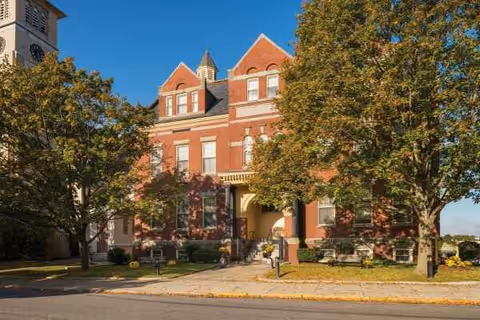 Red brick assisted living building with trees framing a central entrance along a quiet street.