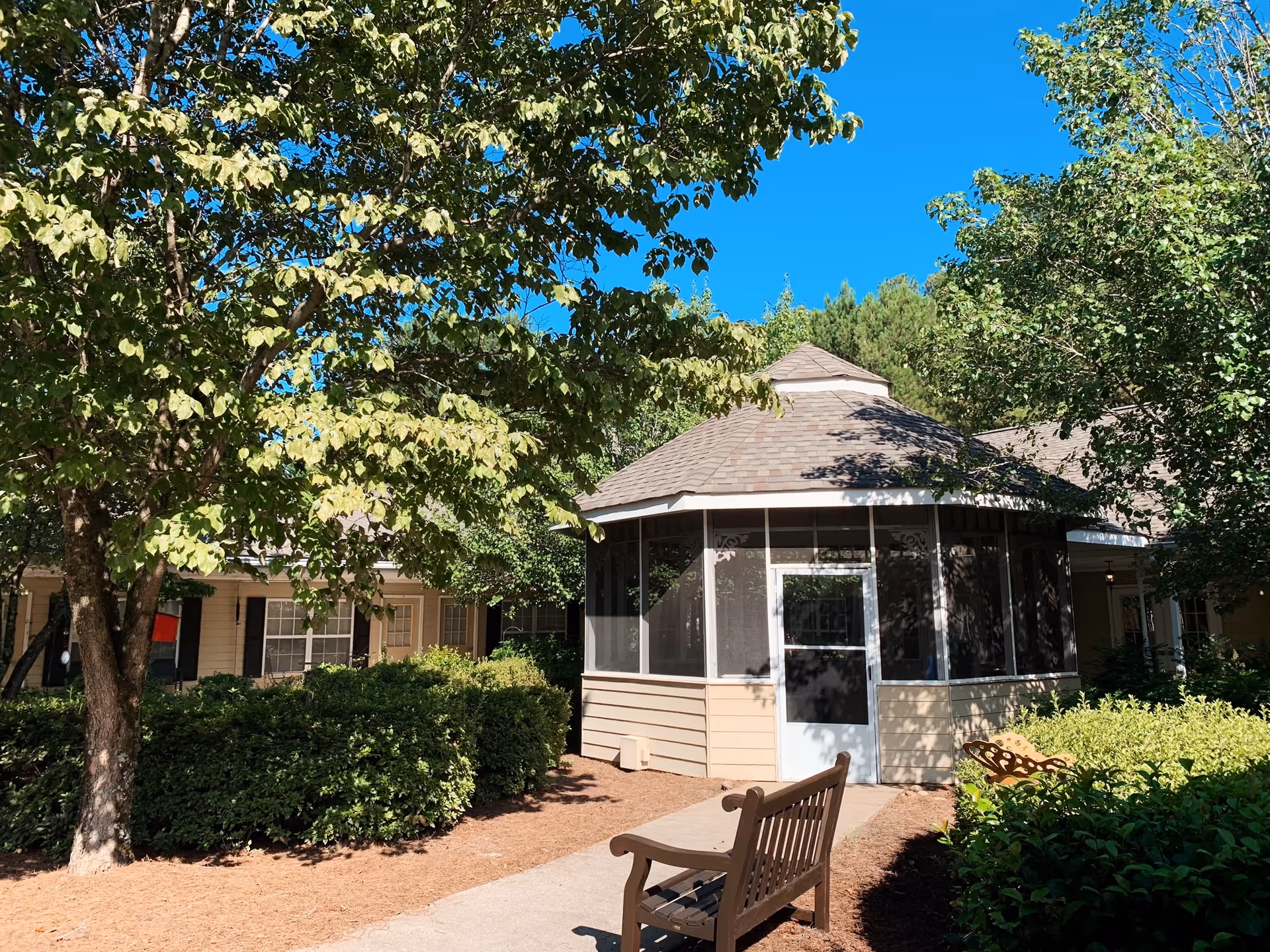 Outdoor view of a senior living facility courtyard with a screened gazebo surrounded by trees and bushes. A wooden bench is placed on a paved pathway leading to the gazebo entrance under a clear blue sky.