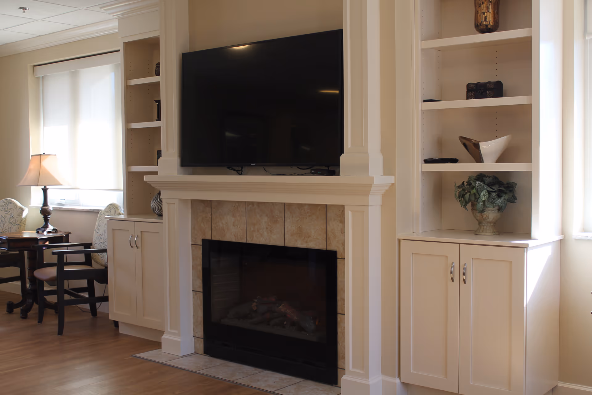 A cozy living room area featuring a wall-mounted flat-screen TV above a tiled fireplace. On either side of the fireplace are built-in white cabinets and shelves decorated with small plants and decorative items. To the left, there is a window with a white roller shade, a table lamp, and two chairs with patterned upholstery. The room has wooden flooring and soft natural lighting.