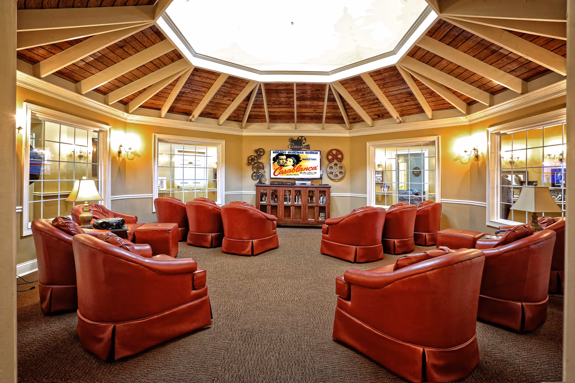 A cozy senior living common area with red leather armchairs arranged in a semicircle facing a TV screen displaying a Casablanca movie poster. The room has a high wooden ceiling with a skylight, beige walls with white trim, and windows on three sides. There are decorative film reels on the wall above a wooden cabinet beneath the TV, and table lamps providing warm lighting.