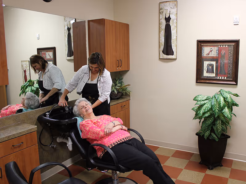 An elderly woman reclines in a salon chair with her eyes closed while a caregiver washes her hair at a sink in a well-lit room with wooden cabinets, a large mirror, framed artwork on the walls, and a potted plant in the corner.