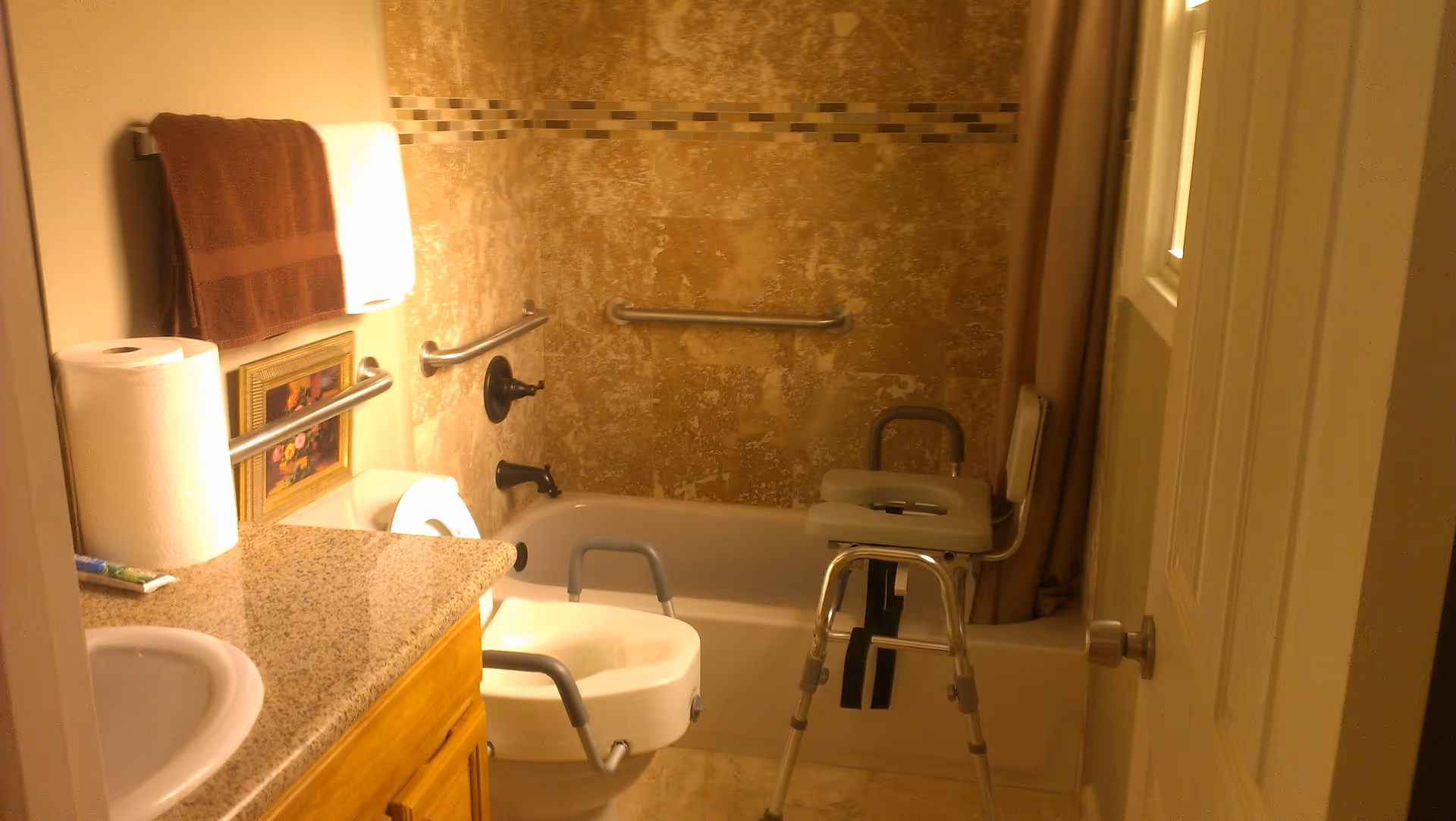 Bathroom with a bathtub equipped with grab bars and a shower chair. There is a sink with a granite countertop, a roll of paper towels, a toothbrush, and a towel rack with brown and white towels. The walls have beige tiles and a decorative tile border.