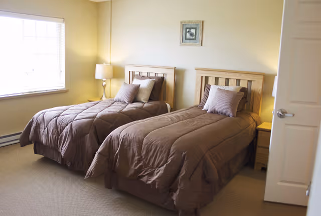 Simple bedroom with two twin beds dressed in brown comforters, a nightstand with a lamp between them and a window on the left.