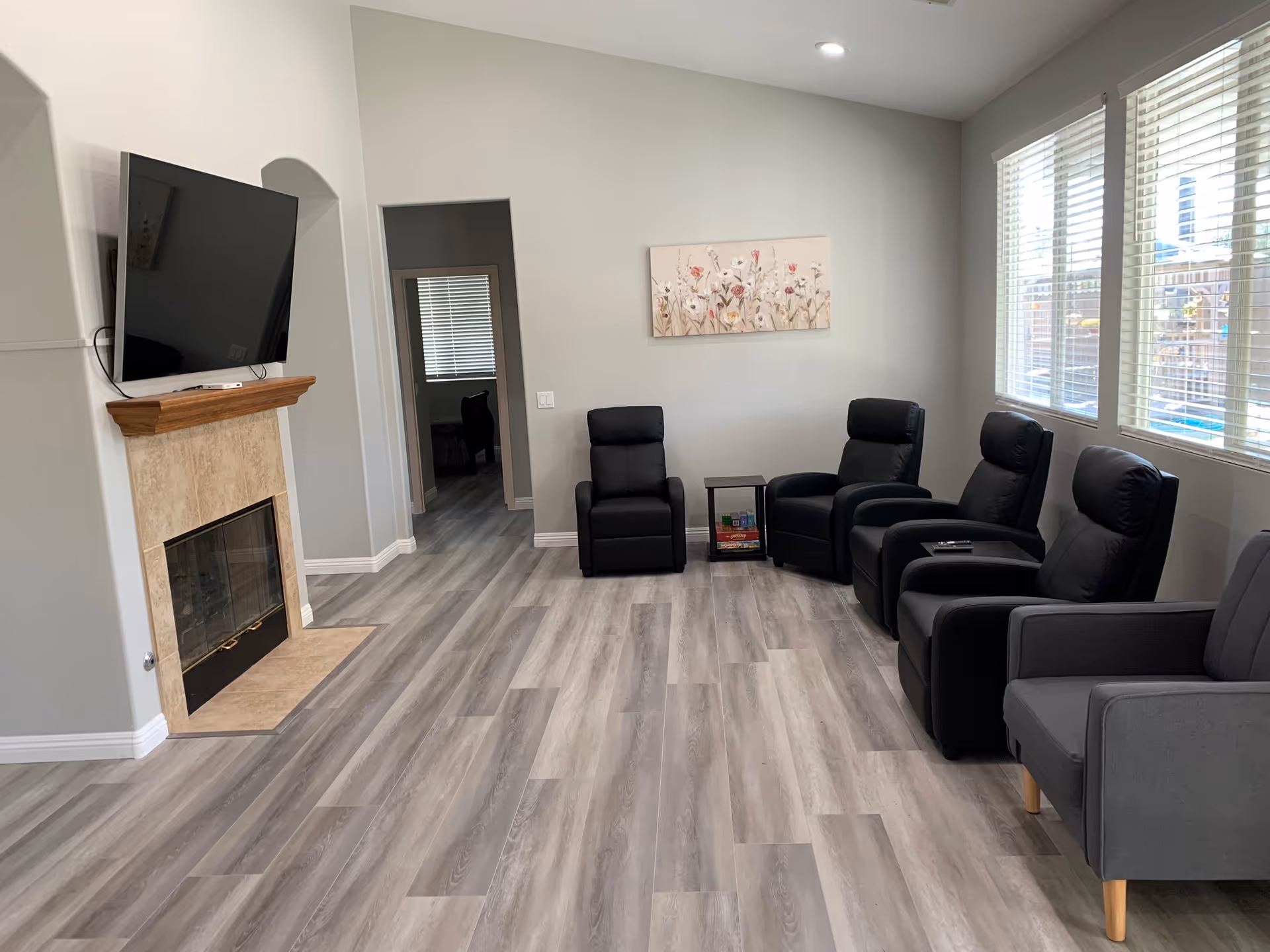 A modern living room with light gray wood flooring, a wall-mounted flat-screen TV above a fireplace with a wooden mantle, and six black and gray armchairs arranged along the walls. A small side table with magazines is placed between two chairs. Large windows with white blinds allow natural light to fill the room, and a floral painting hangs on the wall.