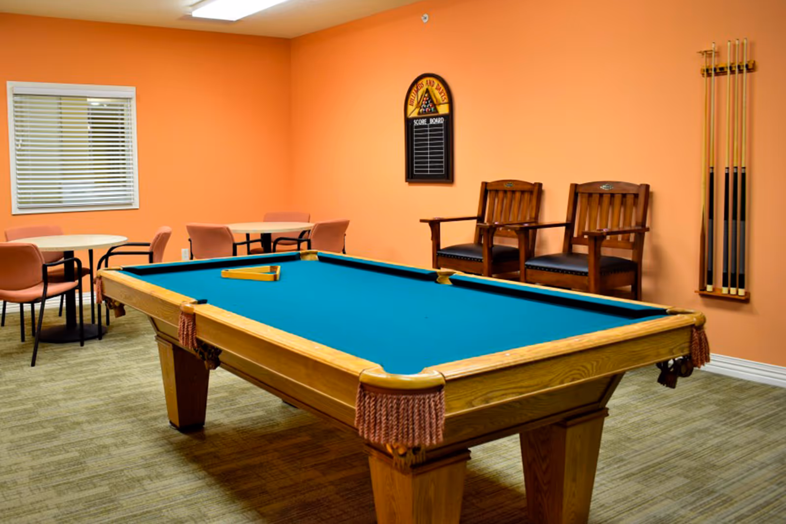 A recreational room with a pool table in the center, two wooden chairs against an orange wall, a rack holding pool cues, and two round tables with pink chairs near a window with blinds.