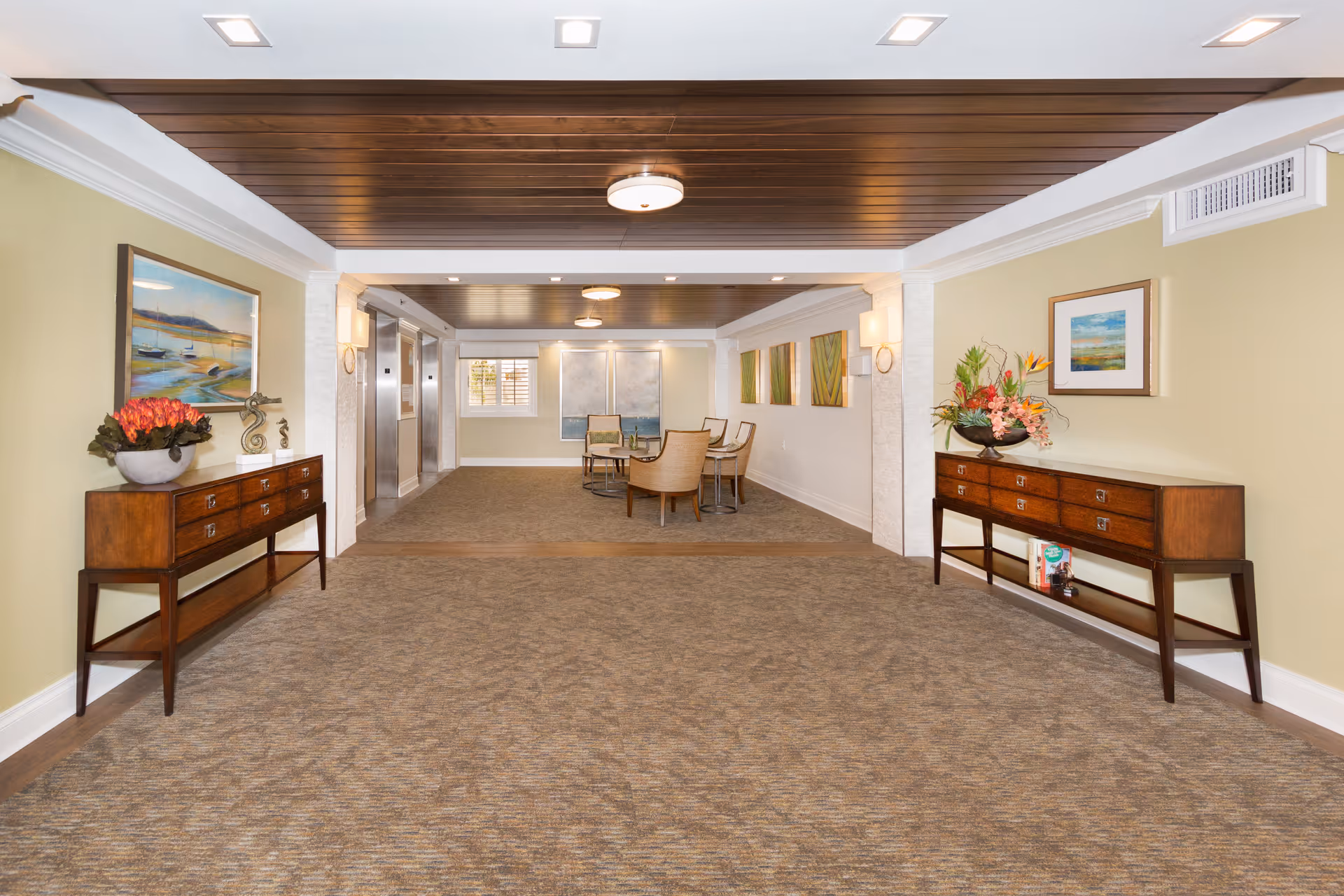 Spacious furnished lobby/common area with carpeted floor, wooden ceiling accents, side console tables with floral arrangements, a seating area and elevators in the background.