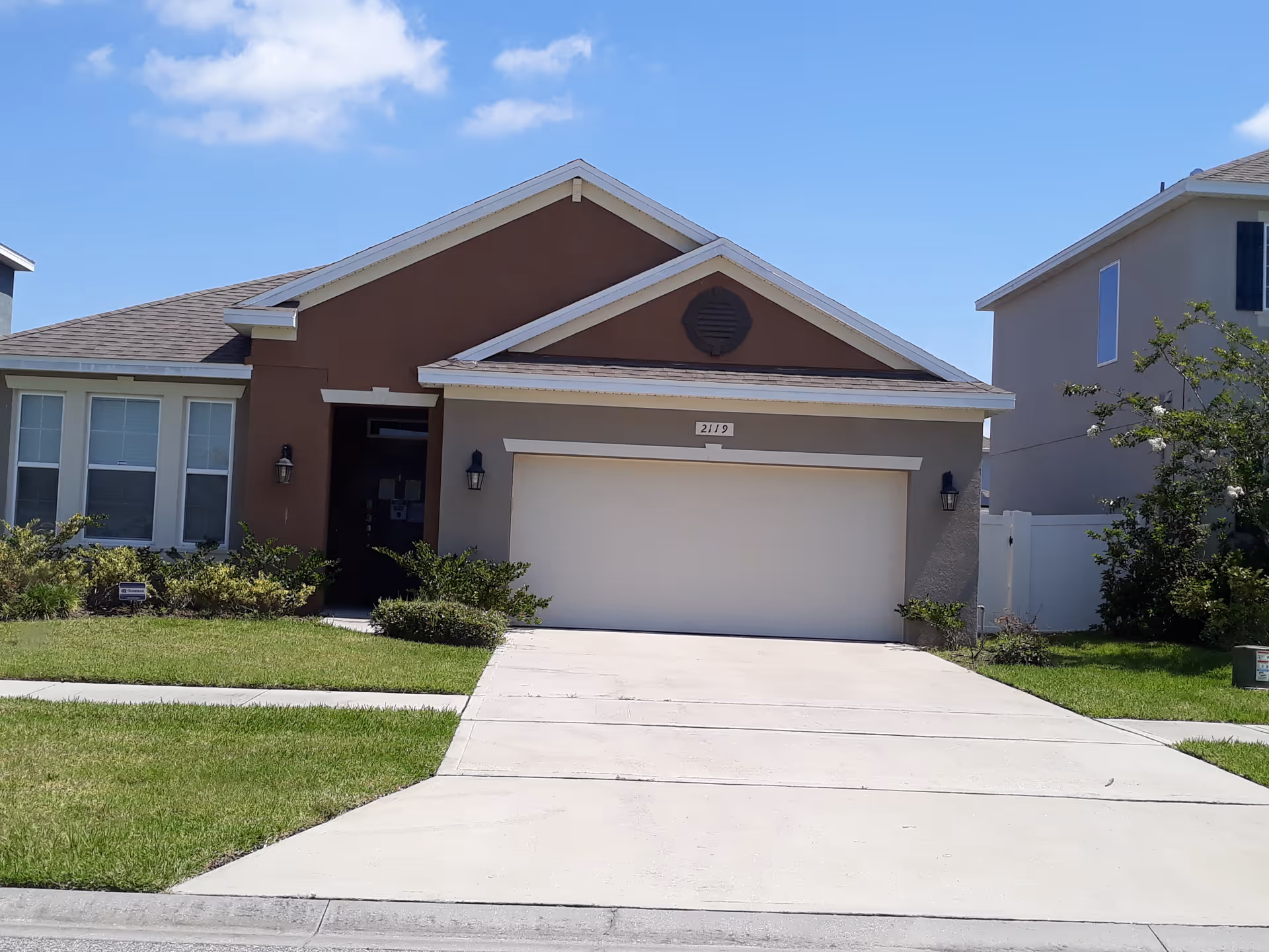 Front exterior of a single-story house with a double garage, driveway, lawn, and neighboring homes under a blue sky.