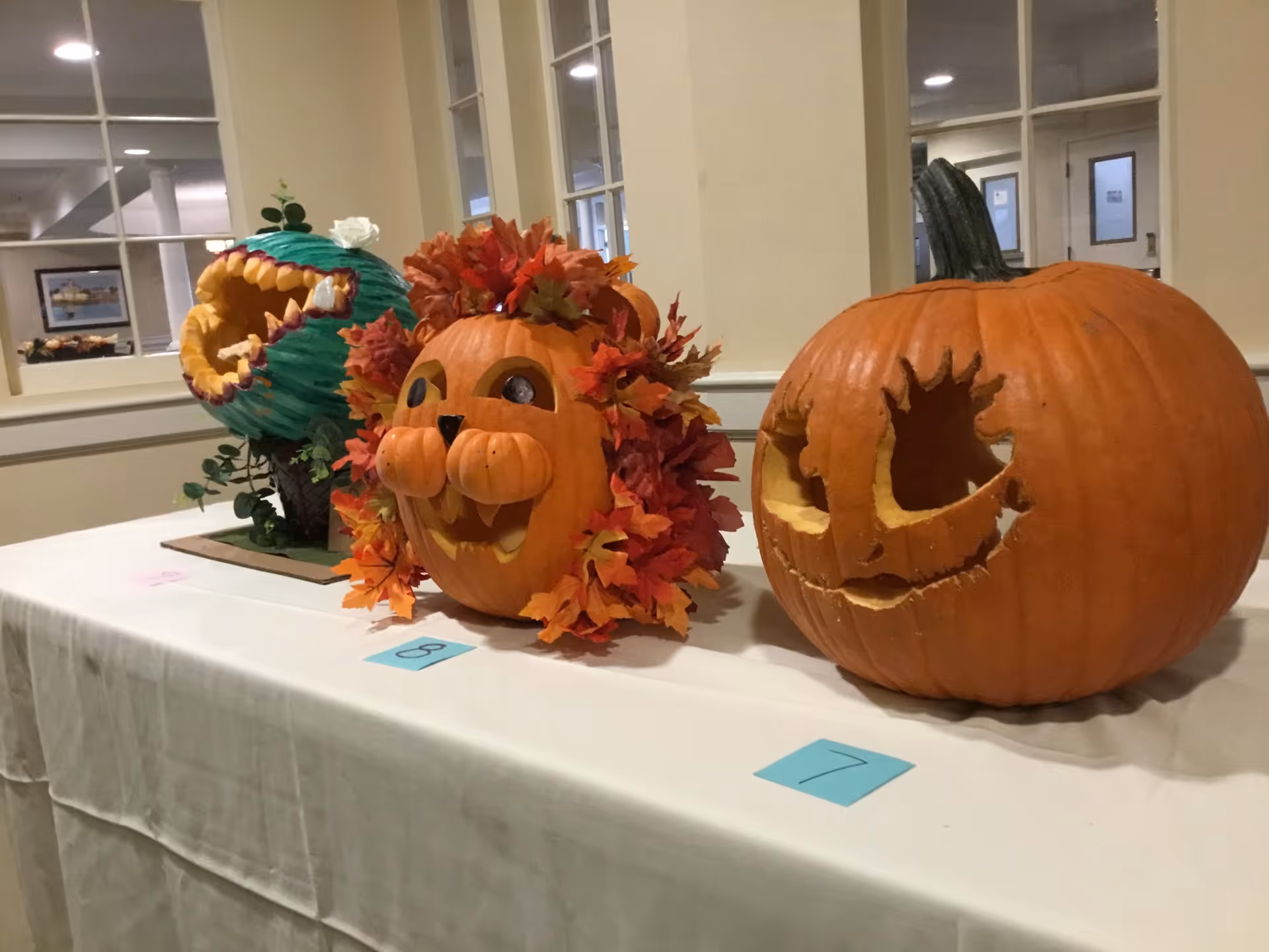 Three creatively carved pumpkins displayed on a white table indoors. The pumpkin on the left is painted green and carved to look like a creature with sharp teeth. The middle pumpkin is carved and decorated with orange and red leaves to resemble a lion's face. The pumpkin on the right has a simple carved face with large eyes and a mouth. Each pumpkin has a numbered card in front of it.