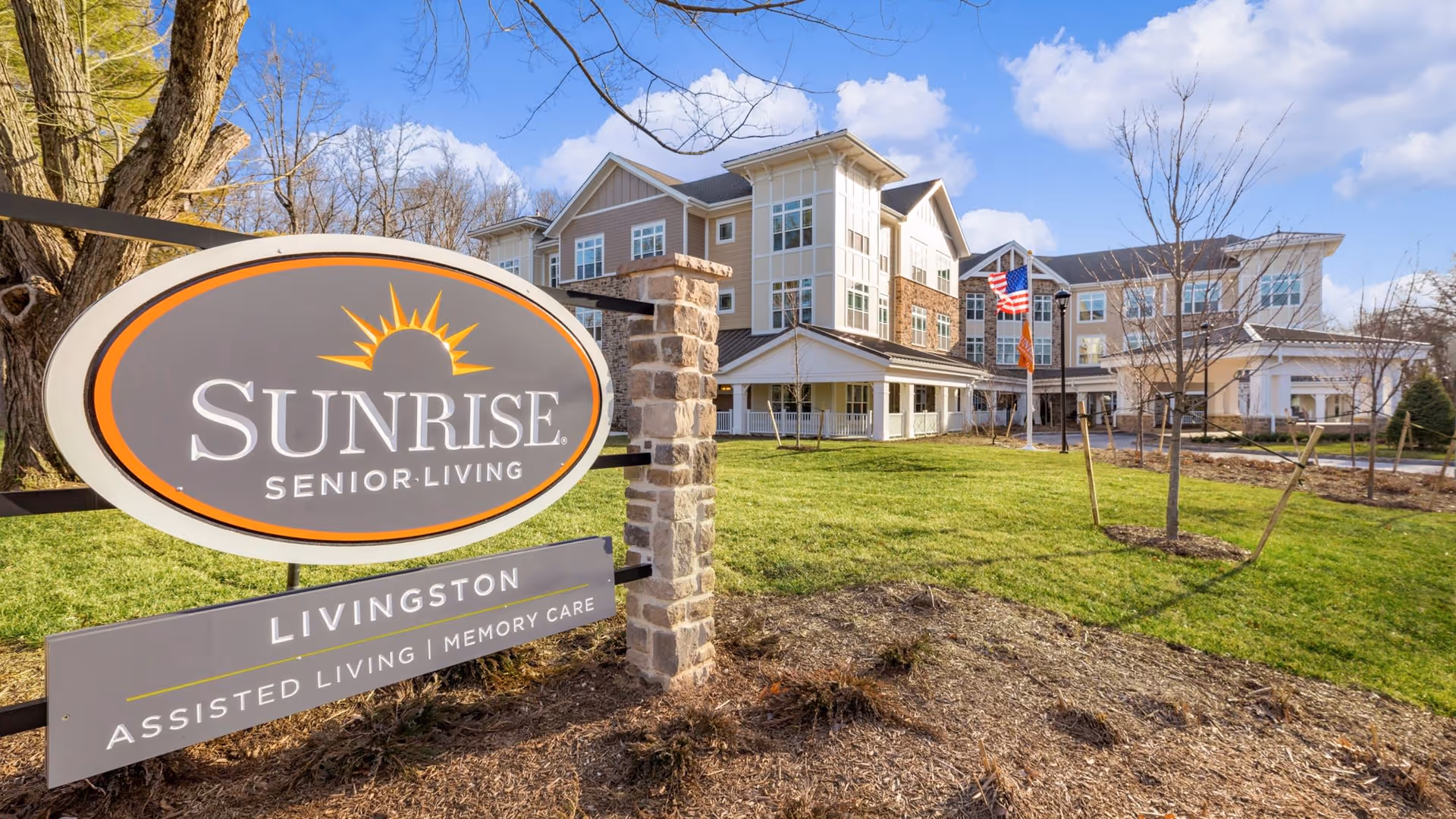 Exterior front of the Sunrise Senior Living Livingston building with a large entrance sign, lawn, and American flag.