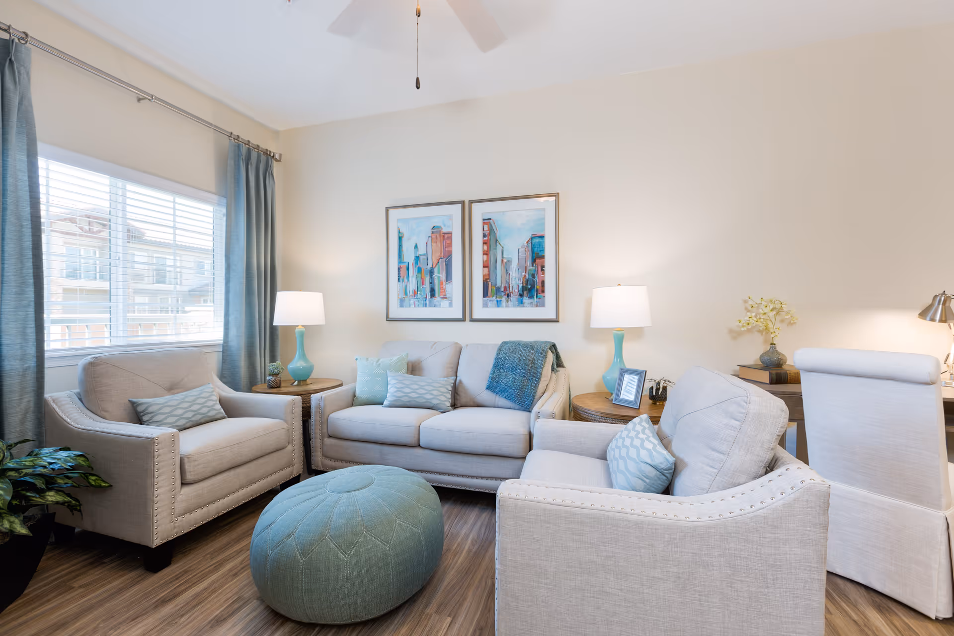 A bright and cozy living room with beige walls and wood flooring. The room features a beige sofa and two matching armchairs with light blue patterned pillows. A round, light blue ottoman is placed in the center. Two framed cityscape paintings hang on the wall above the sofa. There are two side tables with turquoise lamps and decorative items. A window with blue curtains allows natural light to enter the room. A white upholstered chair and a desk with a lamp and flowers are visible on the right side.