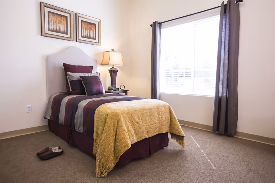 A bright single bedroom with a made twin bed dressed in striped bedding, a bedside table and lamp next to a large window.