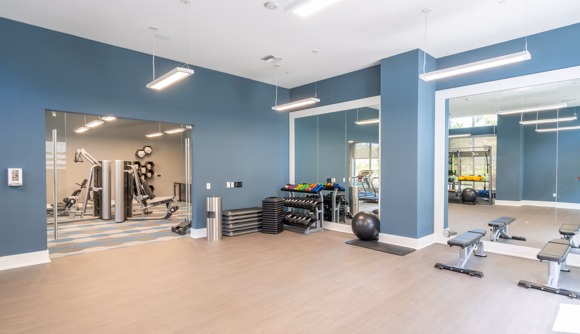 A modern fitness room with blue walls and light wood flooring. The room features large mirrors on two walls, a rack of colorful dumbbells, stacked exercise steps, a black exercise ball on a mat, and two workout benches. Through a glass doorway, additional gym equipment is visible in an adjacent room.
