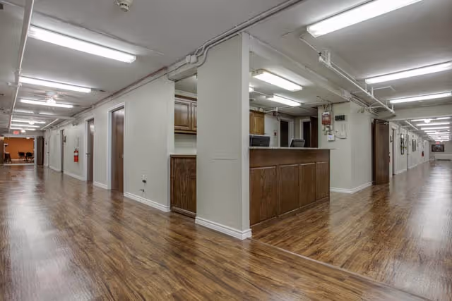 Interior hallway of a senior living facility with wood flooring, beige walls, and fluorescent ceiling lights. There is a reception desk with wood paneling in the center, and doors along the hallway. A fire extinguisher is mounted on the wall to the left.
