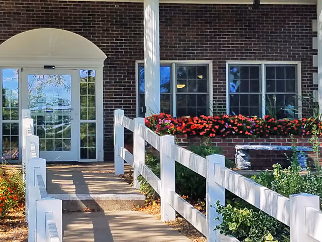 Brick building entrance with a concrete ramp and white railing, double glass doors, windows, and flower beds.