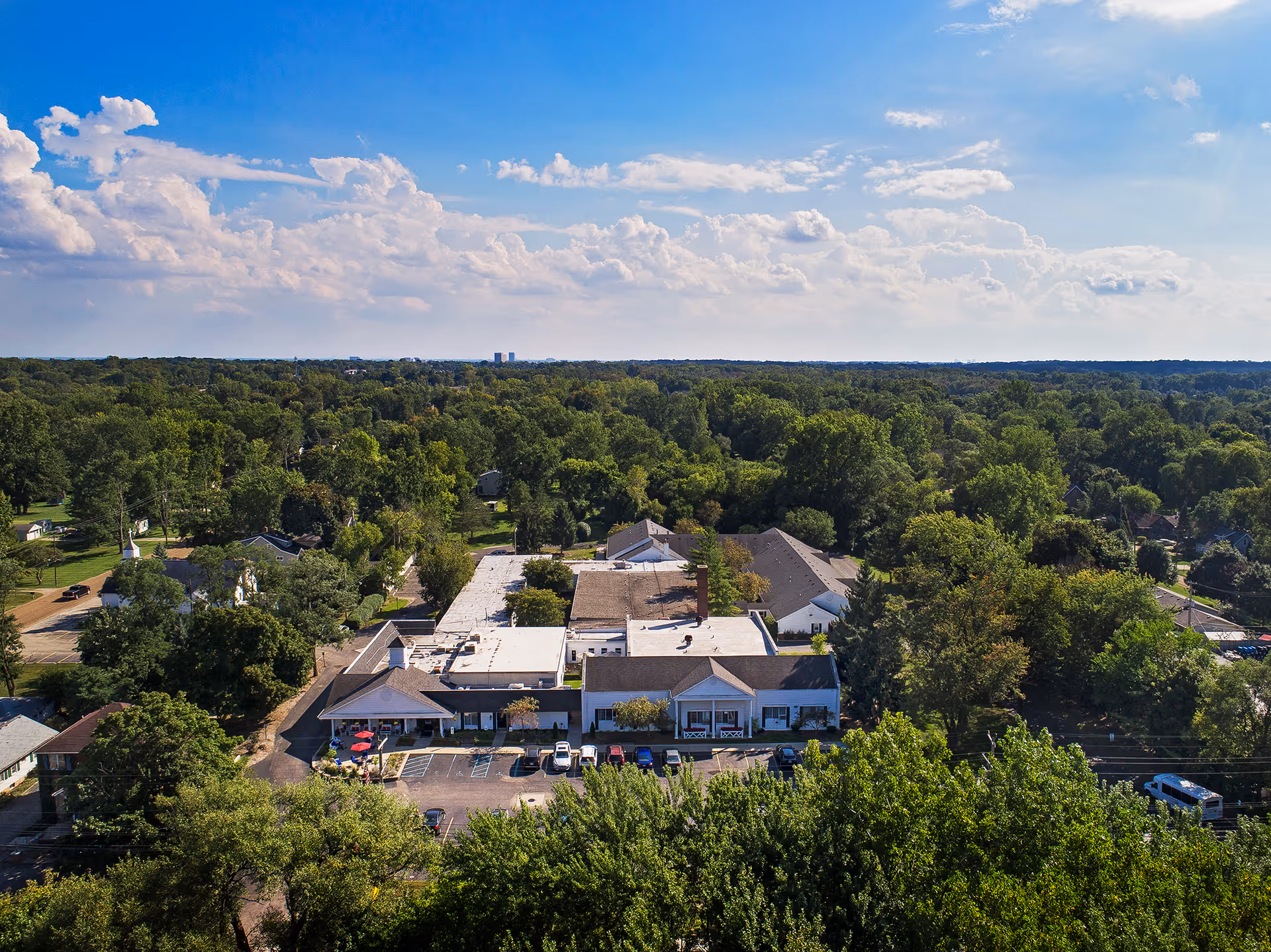 Aerial view of American House Elmwood facility surrounded by dense green trees under a partly cloudy blue sky, with a parking lot and several cars visible in front of the building.