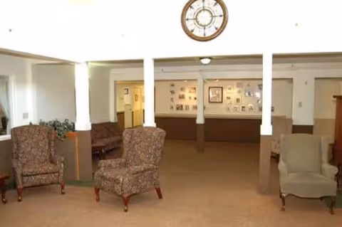 A spacious common area with beige carpet and white walls, featuring three upholstered armchairs arranged near white pillars. A large round clock is mounted on the wall above the pillars, and framed pictures are displayed on the far wall.