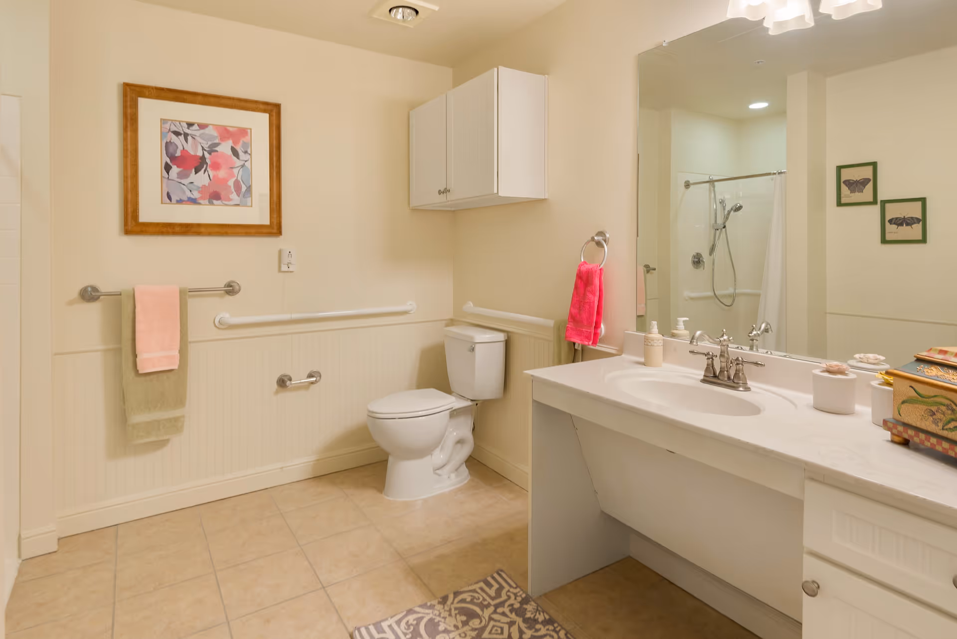 A clean and well-lit bathroom with a toilet, a large vanity with a sink, a mirror, and a shower with a curtain. The walls are light-colored with a framed floral artwork and two butterfly pictures. There are towels hanging on rails and a cabinet mounted on the wall above the toilet.
