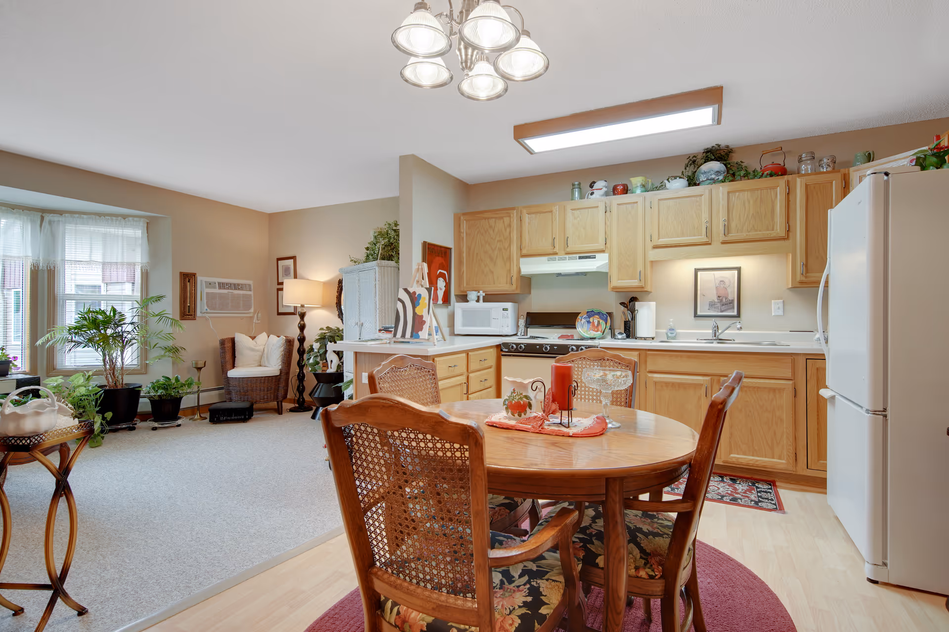 A cozy kitchen and dining area in a senior living facility with wooden cabinets, a white refrigerator, microwave, stove, and sink. A round wooden dining table with four chairs is set on a red rug, and the adjacent living area has a wicker chair, plants, and a window with white curtains.