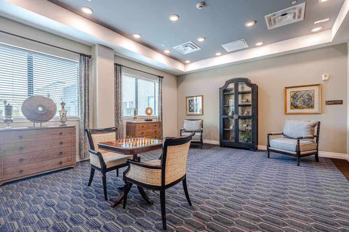 Well-lit communal lounge with a chessboard table, upholstered chairs, display cabinets, and large windows.
