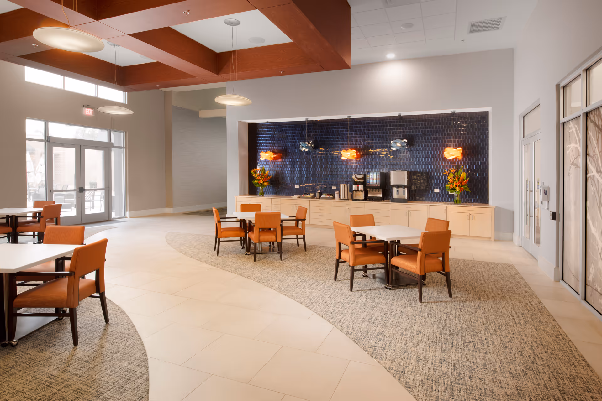 A spacious dining area with several tables and orange cushioned chairs arranged on a carpeted and tiled floor. The back wall features a coffee and beverage station with a dark blue tiled backsplash and decorative hanging lights. Large windows and glass doors allow natural light to fill the room.