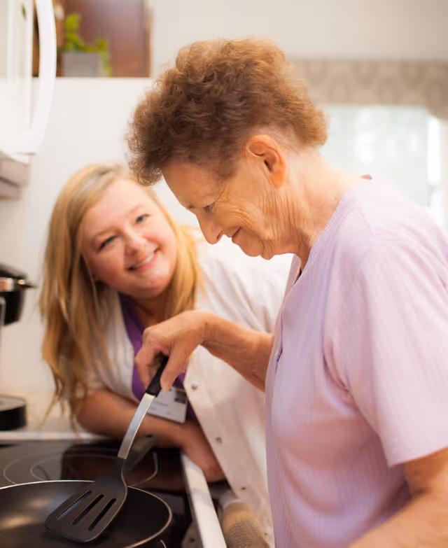 An elderly woman cooking with a spatula in a kitchen while a younger woman, possibly a caregiver or nurse, smiles and watches her.