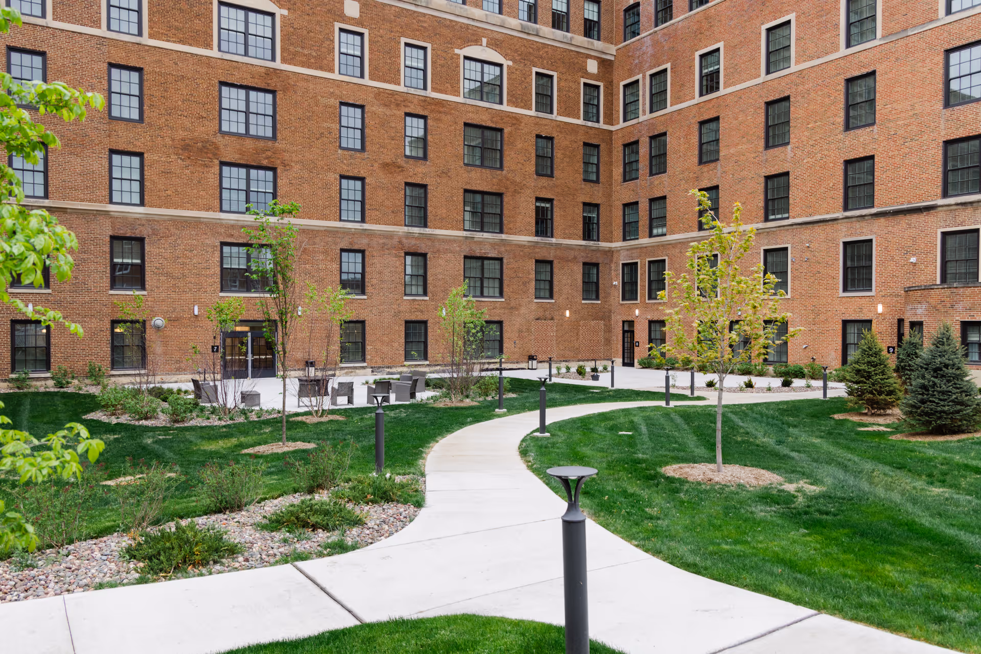Outdoor courtyard area with a curved concrete pathway surrounded by green grass, small trees, and landscaped plants. The courtyard is enclosed by a multi-story brick building with many windows.