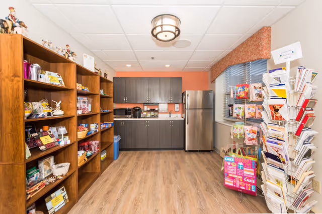 Small kitchenette and market area with wooden shelves of snacks on the left, a greeting-card rack on the right, and cabinets with a stainless refrigerator at the back.