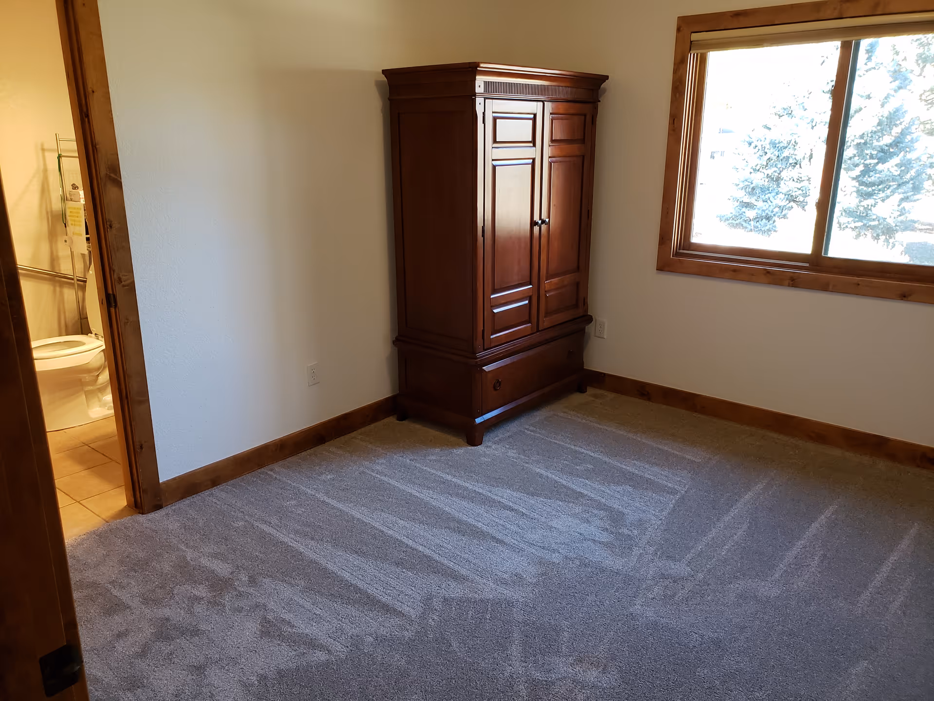 Empty carpeted bedroom with a wooden armoire, a window, and a doorway leading to a bathroom.