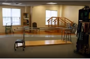 A well-lit physical therapy room with parallel bars and balance beams, walkers, stools, and exercise equipment near windows.