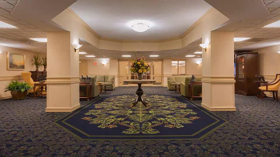 A spacious, well-lit sitting area in a senior living facility with patterned blue and green carpet, beige walls, and a central table holding a large floral arrangement. The room features multiple seating areas with green upholstered chairs and wooden side tables, decorative plants, and framed artwork on the walls.